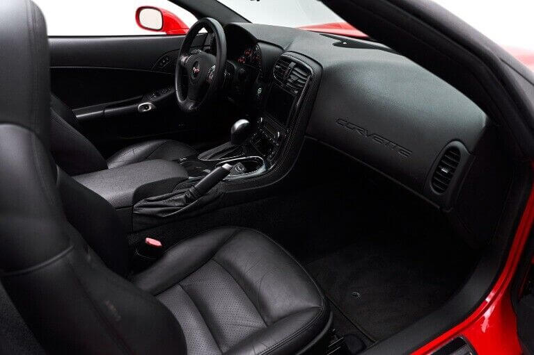 Corvette C6 Seat Covers - Black - Lux Seat CoversInterior view of a Corvette C6 featuring black faux leather seat covers on both front seats, showcasing the sleek and snug fit of the covers, detailed stitching, and ergonomic design. The red interior accents and dashboard provide a striking contrast, highlighting the high-quality material and fit of the seat covers.